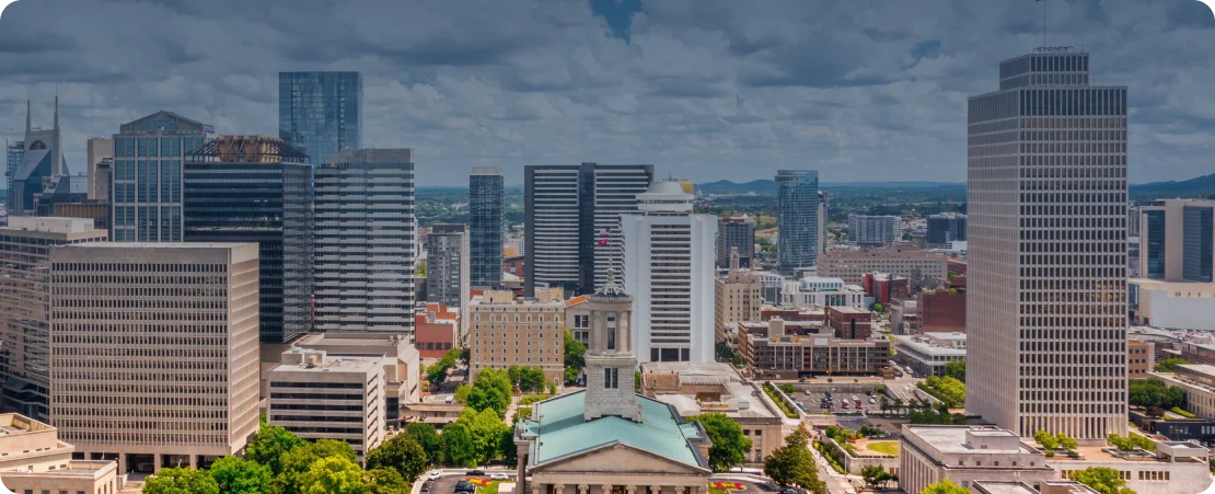 Aerial view of a cityscape with mid-rise and high-rise buildings, including a prominent historic building with a green roof in the foreground, under a partly cloudy sky
