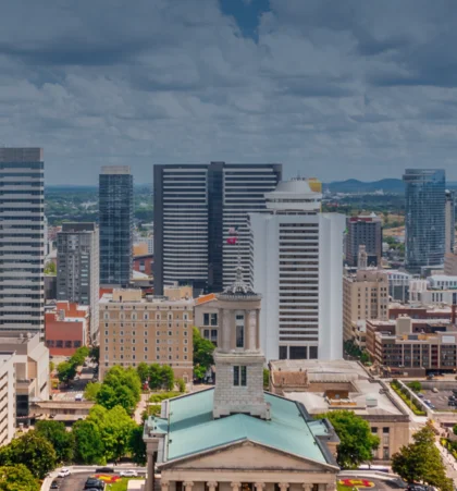 Aerial view of a cityscape with mid-rise and high-rise buildings, including a prominent historic building with a green roof in the foreground, under a partly cloudy sky